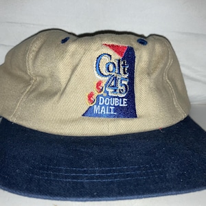 May include: Tan and navy blue baseball cap with the embroidered text "Colt 45 Double Malt". The cap has a curved brim and a six-panel design. The logo is in blue, red, and white.