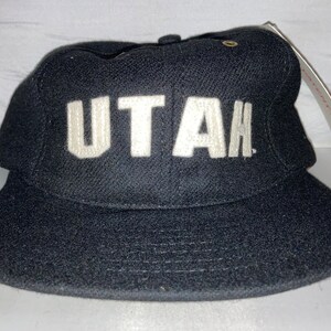 May include: A black baseball cap with a white "UTAH" embroidered on the front. The cap has a curved brim and a white tag attached to the side.