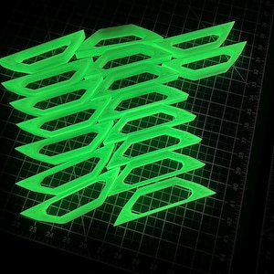 May include: A stack of neon green, geometric shapes with a glowing effect. The shapes are angular with a rectangular center and pointed ends. The objects are arranged on a dark surface with measurement markings.