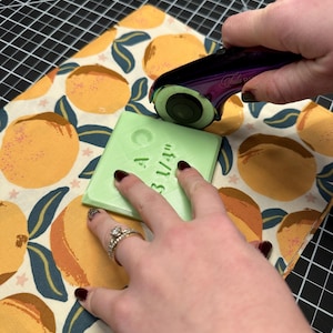Puede incluir: Un cortador rotatorio con mango morado se utiliza para cortar un cuadrado verde con el texto "A 3 1/4" sobre una tela con estampado de cítricos. La tela está sobre una esterilla de corte negra.