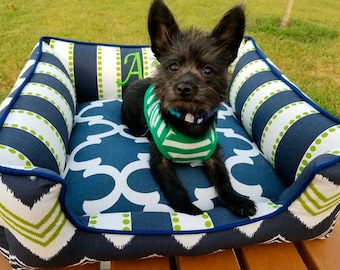 May include: A small black dog with a green and white striped shirt is lying in a blue and green dog bed. The bed has a white and blue patterned fabric with green polka dots. The bed has a navy blue border and the letter "A" is embroidered on the side.