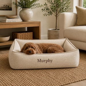 May include: A rectangular, beige fabric pet bed with high, structured sides and a flat base. The front of the bed features the name Murphy embroidered in dark brown thread. A brown, curly-haired dog is resting inside the bed on the cushioned surface.