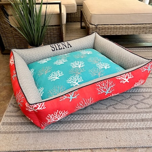 May include: A rectangular pet bed featuring a vibrant coral-colored exterior with a white coral reef pattern. The interior cushion is turquoise with a white coral print. The inner side wall is grey with a small pattern and the name SIENA embroidered.