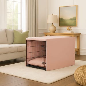 Puede incluir: Un juego de funda y cama para jaula de perro rosa. La jaula es de metal negro con una funda rosa a juego y un cojín rosa en el interior. La jaula está sobre una alfombra color crema en un salón.