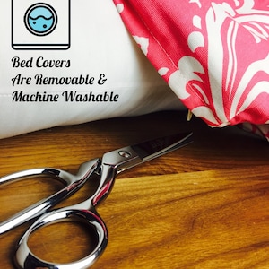 Pu&ograve; includere: Un cuscino bianco con un tessuto drappeggiato sopra, a motivi rosa e bianchi. Il cuscino ha un'icona di lavatrice e il testo "Bed Covers Are Removable & Machine Washable". Un paio di forbici argentate sono su una superficie di legno.