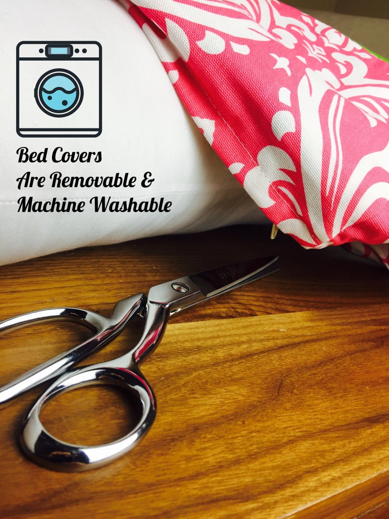 May include: A white pillow with pink and white patterned fabric folded over the top. The pillow has a washing machine icon and the text "Bed Covers Are Removable & Machine Washable". A pair of silver scissors are on a wooden surface.