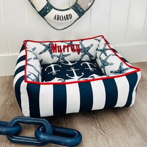 Peut inclure: Un lit pour chien rayé blanc et bleu marine avec une bordure rouge. Le lit a un coussin amovible avec un fond bleu marine et des ancres blanches. Le coussin a également un nom brodé en rouge "Murray" et un motif d'étoile de mer.