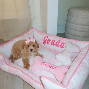 May include: A pink and white dog bed with a bone-shaped pillow. The bed is embroidered with the name "Gouda". A small brown dog with a pink bow is lying on the bed.