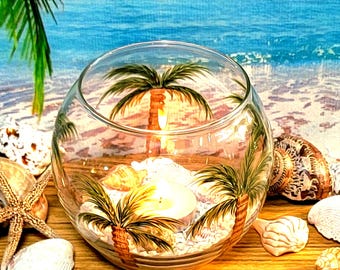 Op de afbeelding: Een glazen kom als kaarshouder met geschilderde palmbomen aan de buitenkant. In de kom liggen wit zand en kleine schelpen rondom een wit waxinelichtje. De kom staat tussen diverse schelpen en een zeester op een ondergrond.