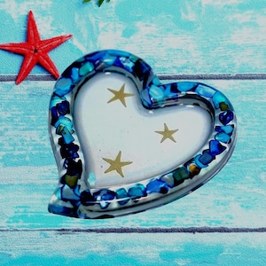 May include: A heart-shaped picture frame with a clear border embedded with blue and turquoise stones. The frame's interior is white with three gold starfish designs. A red starfish rests beside the frame on a blue wooden surface.