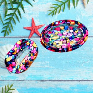 May include: Two decorative soap dishes with colorful mosaic designs. One is oval-shaped, and the other is a smaller, kidney-shaped dish. A red starfish and seashells are also visible on a blue wooden surface.