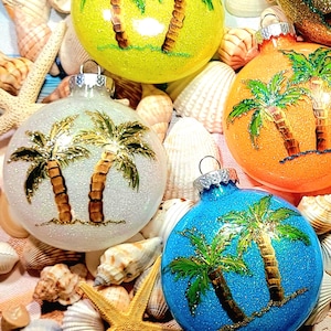 May include: Four round Christmas ornaments with a beach theme. Each ornament features two palm trees with green leaves and gold trunks on a glittery background. The ornaments are white, yellow, orange, and blue.