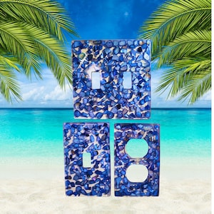 May include: Three rectangular wall plates featuring a mosaic of small, iridescent blue shell fragments embedded in a clear resin base. The set includes one double toggle switch plate, one single toggle switch plate, and one duplex outlet cover plate.