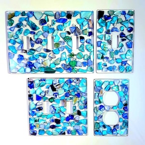 May include: Set of four decorative light switch covers and outlet covers. The covers are adorned with a mosaic of blue, teal, and green glass pieces. The set includes covers for three switches, one switch, two switches, and a double outlet.