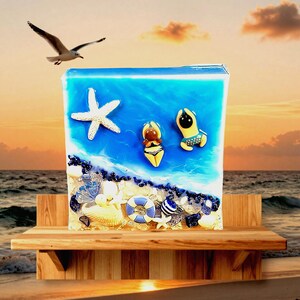May include: A beach-themed resin art piece featuring a blue ocean scene with a starfish, two figures in swimwear, and seashells. A seagull flies in the background. The artwork is displayed on a wooden shelf, evoking a coastal vibe.
