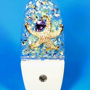 May include: A decorative night light with a white base and a clear, glitter-filled top. A gold starfish with purple and pink accents is the focal point. The night light is designed to plug into a standard wall outlet.