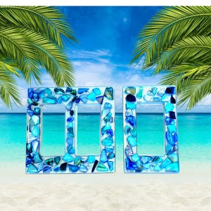 May include: Two decorative light switch covers with a mosaic design. The covers are rectangular with two openings and are adorned with blue and turquoise glass pieces. The background features a beach scene with palm trees and a blue sky.