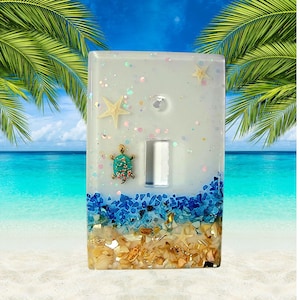 May include: Decorative light switch cover with a beach theme. The cover is clear with embedded seashells, blue glass, and a turquoise turtle. A starfish and glitter add to the coastal design. The switch plate is rectangular.