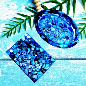 May include: Two blue decorative items, one oval and one rectangular, are filled with small, irregular blue and white pieces. The items are on a light blue surface with green leaves and a wooden handle.
