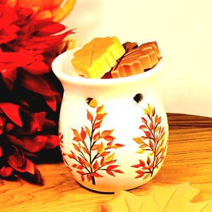 May include: A white ceramic wax warmer with a leaf design in orange, yellow, and red. The warmer contains two leaf-shaped wax melts in shades of yellow and brown. The warmer sits on a wooden surface, with a red floral arrangement in the background.