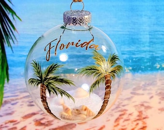 Puede incluir: Un adorno de Navidad de vidrio transparente con una escena pintada de palmeras y conchas. El adorno tiene la palabra "Florida" escrita en letras doradas.