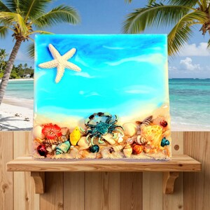 May include: A square resin art piece depicting a beach scene with a starfish, crab, seashells, and sand. The background features a blue sky and ocean. The artwork is displayed on a wooden shelf.