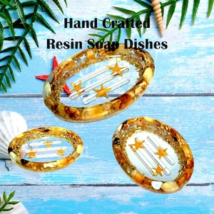 May include: Hand-crafted resin soap dishes in a beach theme. The dishes are oval-shaped with a clear resin base, embedded with gold and brown flecks. Each dish contains three clear, parallel bars and gold star accents. The background is a blue and white wood-grain pattern.