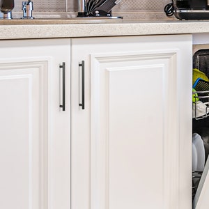 May include: Two white cabinet doors with black metal bar handles. The cabinet doors are closed and have a raised panel design.