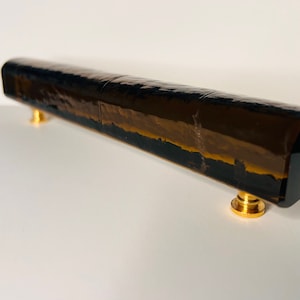 Może przedstawiać: Długi, ciemnobrązowy szklany uchwyt do szafki z złotymi metalowymi akcentami. Uchwyt ma fakturowaną powierzchnię i prostokątny kształt.
