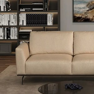 May include: A beige sofa with a wooden frame and black metal legs sits in front of a bookshelf filled with books. A black coffee table with a glass top sits in front of the sofa. A painting hangs on the wall behind the sofa.