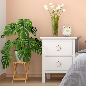 Puede incluir: Mesita de noche blanca con dos cajones y tiradores de anillo dorados. La mesita de noche tiene un despertador blanco en la parte superior y un jarrón de tulipanes blancos. Una planta verde en maceta está en un pequeño soporte de madera a la izquierda de la mesita de noche.
