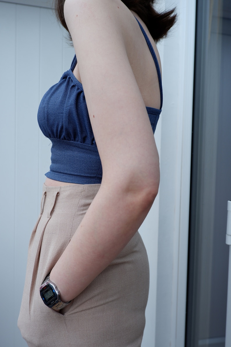 Pu&ograve; includere: Una donna indossa un top corto blu con un paio di pantaloni beige a vita alta. Indossa un orologio digitale al polso sinistro.
