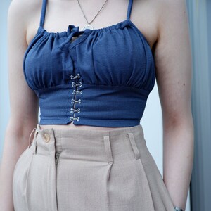 Pu&ograve; includere: Un top corto blu con scollo arricciato e una fila di chiusure in metallo argentato sul davanti. Il top &egrave; abbinato a pantaloni beige a vita alta.