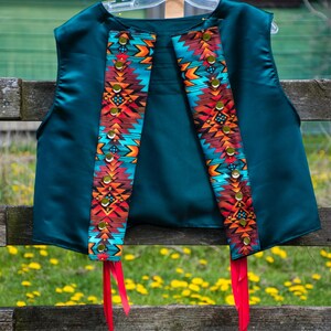 May include: Teal vest with colourful patterned panels and red ribbon ties. The panels feature a geometric design in shades of orange, turquoise, and brown, accented with gold-coloured studs. The vest hangs on a wooden hanger against a rustic fence.
