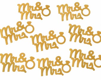Peut inclure: Un ensemble de huit découpes en papier de couleur dorée, chacune présentant le texte "Mr & Mrs" dans une police cursive, accompagné d'une icône d'anneau circulaire avec une petite forme de diamant au sommet, disposées en motif éparpillé.