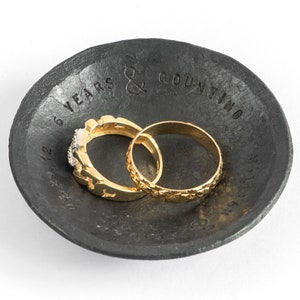 May include: A black metal ring dish with the inscription "6 Years & Counting" and two gold rings inside.