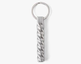 May include: A silver metal keyring with a twisted, textured design. The keyring has a circular ring at the top for attaching keys. The twisted bar is rectangular and has a matte finish. The background is plain white.