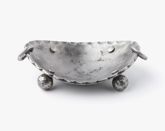 May include: A small, silver-colored metal bowl with a hammered texture. The bowl has a scalloped rim and is supported by two ball-shaped feet. It features two ring handles on either side.