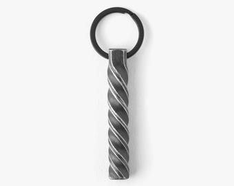 May include: A metal keyring with a twisted, textured design. The keyring has a black ring and a silver-coloured, rectangular pendant with a spiral pattern. The background is plain white.