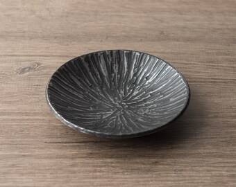 May include: A small, round, dark gray bowl with a textured, radial pattern. The bowl has a rough, almost metallic appearance, and sits on a wooden surface. The bowl is likely used for serving small portions of food or as a decorative item.