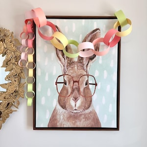 May include: A framed rectangular print featuring a brown rabbit wearing large red-rimmed glasses against a light blue background with white dashes. A decorative paper chain in pastel shades of pink, yellow, and green hangs over the top frame.