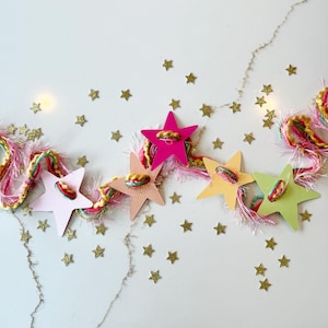 May include: A garland featuring five multi-colored star-shaped cutouts in shades of pink, peach, yellow, and light green, strung together with textured pink and multi-colored yarn, accented by scattered small gold glitter star confetti.