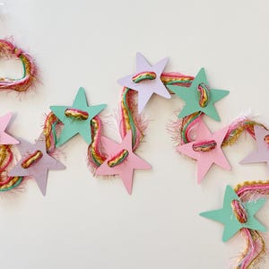 May include: A decorative garland featuring alternating light pink, lavender, and mint green star-shaped cutouts strung together with a multi-colored, textured yarn that includes strands of pink, yellow, green, and metallic gold.