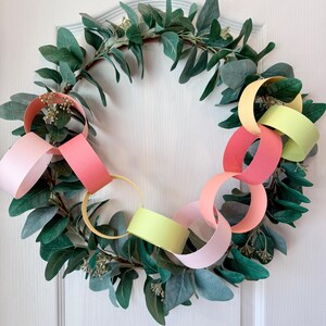 May include: A decorative wreath featuring a ring of green leaves and colorful paper chain links in shades of pink, peach, and yellow. The wreath is hanging on a white door.