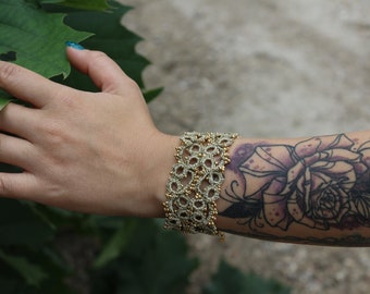 Puede incluir: Un delicado brazalete de ganchillo en oro y beige con un diseño floral. El brazalete se lleva en la muñeca de una persona con un tatuaje de una rosa.