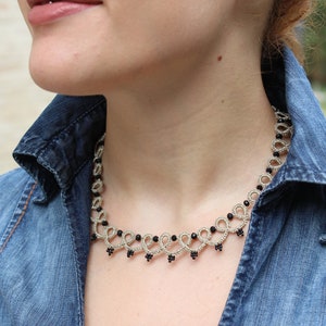 May include: A silver and black beaded necklace with a delicate, intricate design. The necklace is worn by a person wearing a denim shirt.