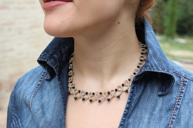 May include: A silver and black beaded necklace with a delicate, intricate design. The necklace is worn by a person wearing a denim shirt.