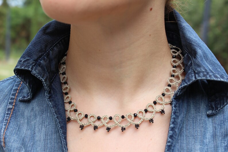 May include: A silver and black beaded necklace with a delicate, intricate design. The necklace is worn by a person with a denim shirt.