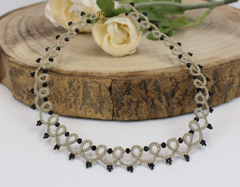 May include: A delicate silver-colored necklace with black beads, featuring a looped design. The necklace is displayed on a wooden surface.