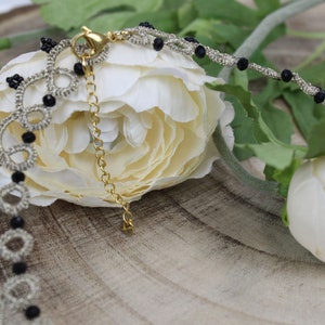 May include: A silver and black beaded necklace with a delicate chain and a clasp. The necklace is lying on a wooden surface with white flowers.